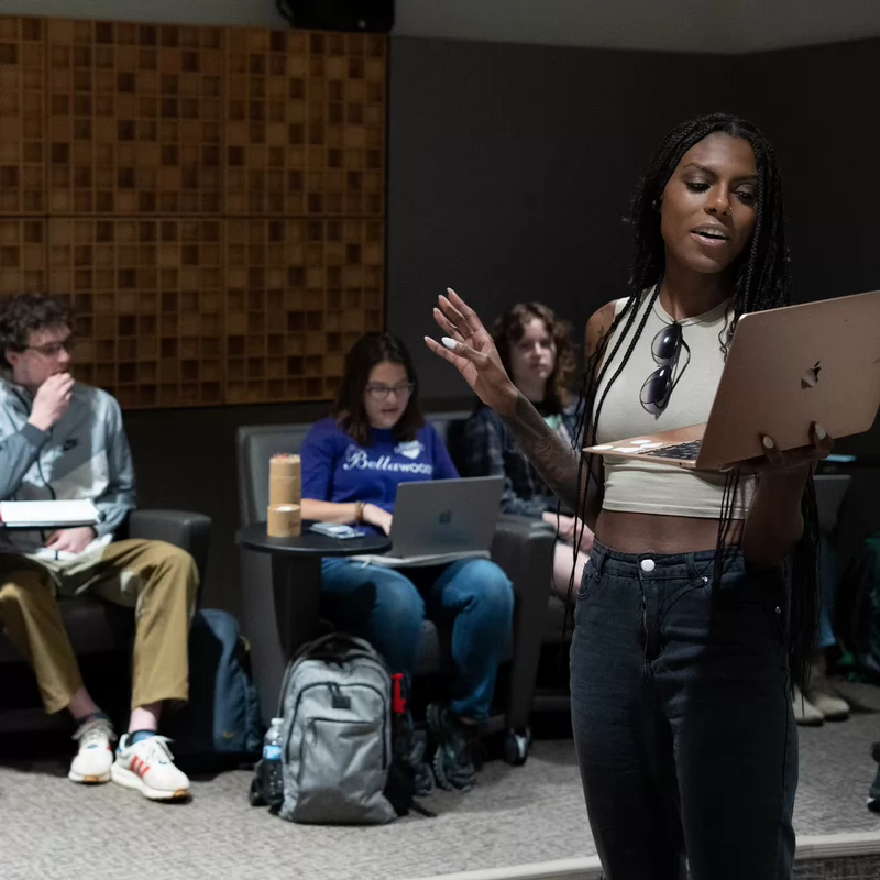 A student stands at the front of a classroom reading from a laptop, speaking to peers seated in cushioned chairs. The room features acoustic paneling on the wall, and other students are engaged with notebooks or laptops during what appears to be a film or TV writing class.