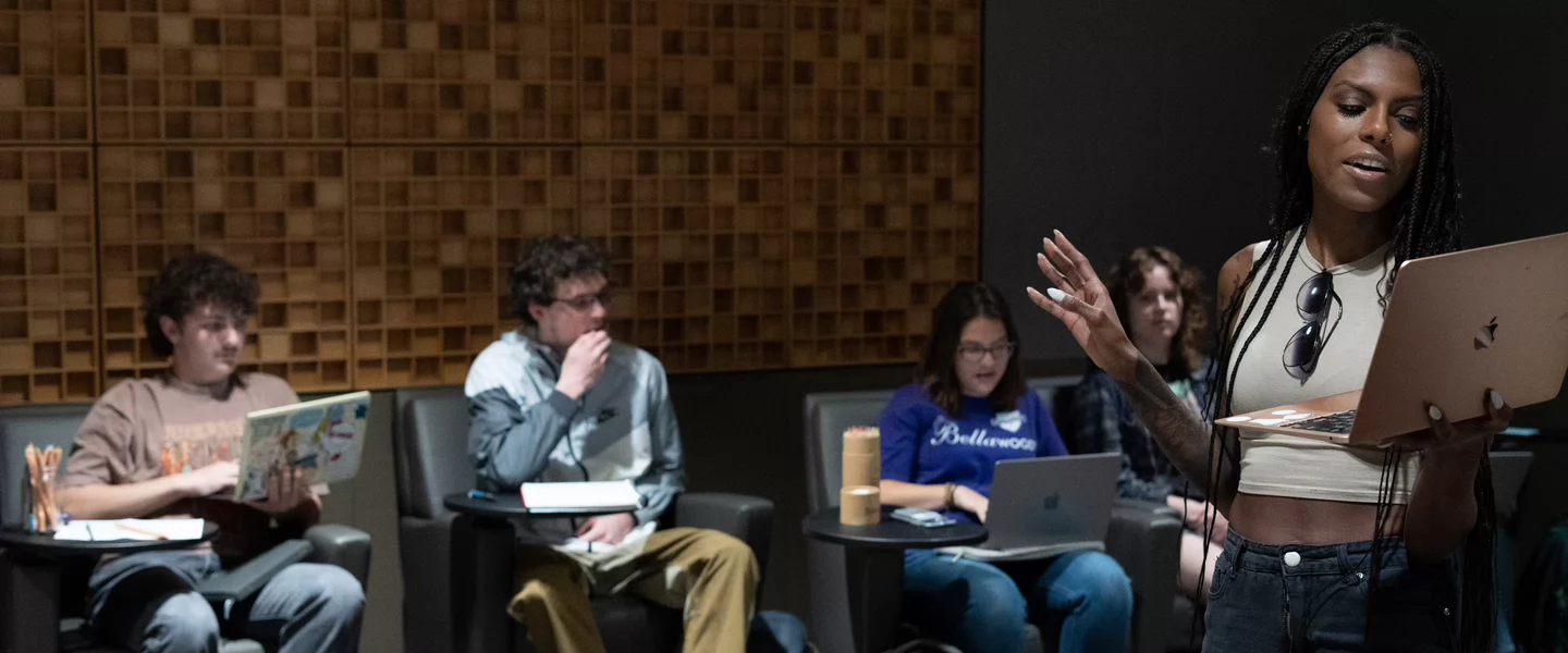 A student stands at the front of a classroom reading from a laptop, speaking to peers seated in cushioned chairs. The room features acoustic paneling on the wall, and other students are engaged with notebooks or laptops during what appears to be a film or TV writing class.