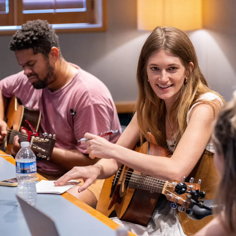 Three students sit around a table in a recording studio, playing guitars, writing notes, and collaborating on songwriting ideas with smiles and focused energy.