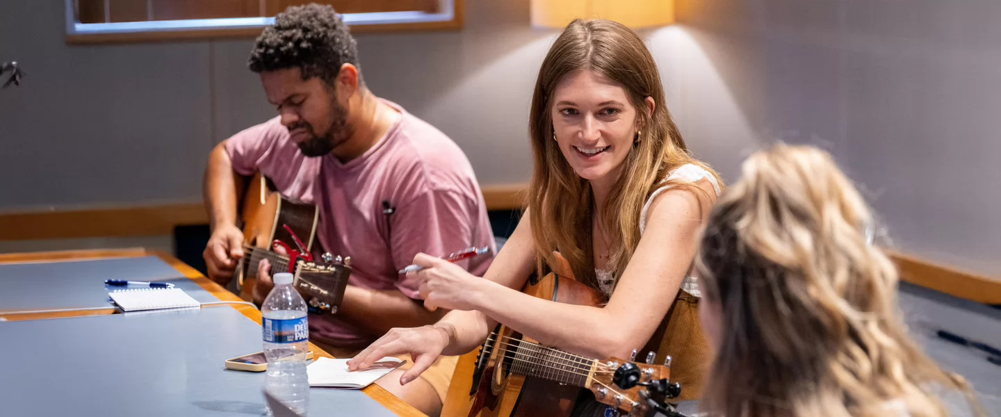 Three students sit around a table in a recording studio, playing guitars, writing notes, and collaborating on songwriting ideas with smiles and focused energy.