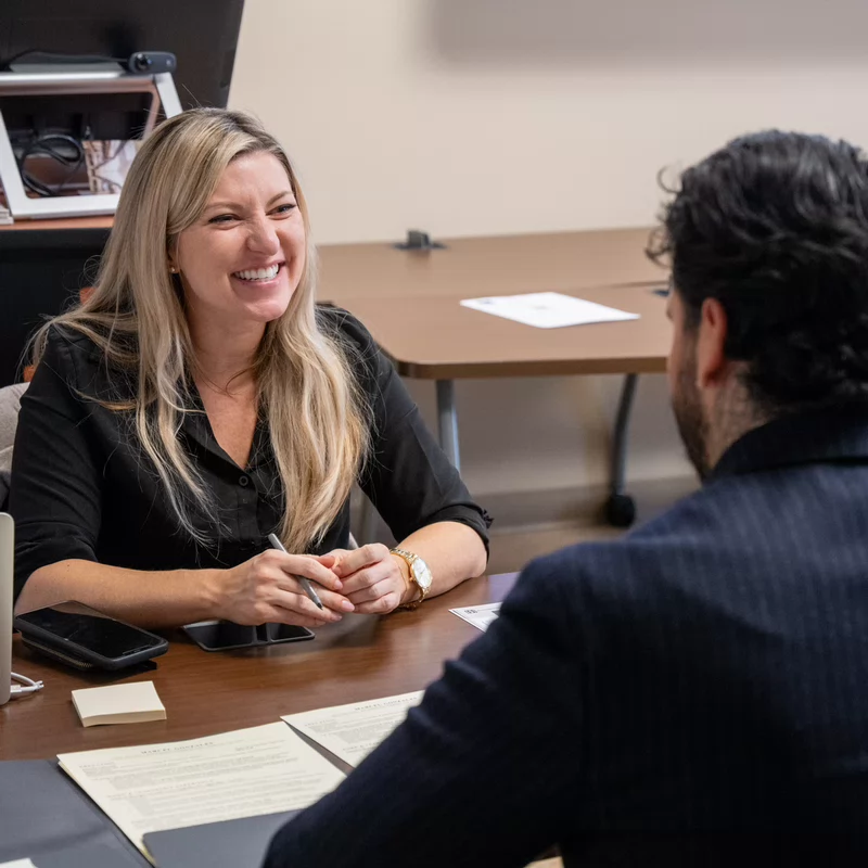 A smiling woman sits at a desk across from another person, engaging in a one-on-one discussion.