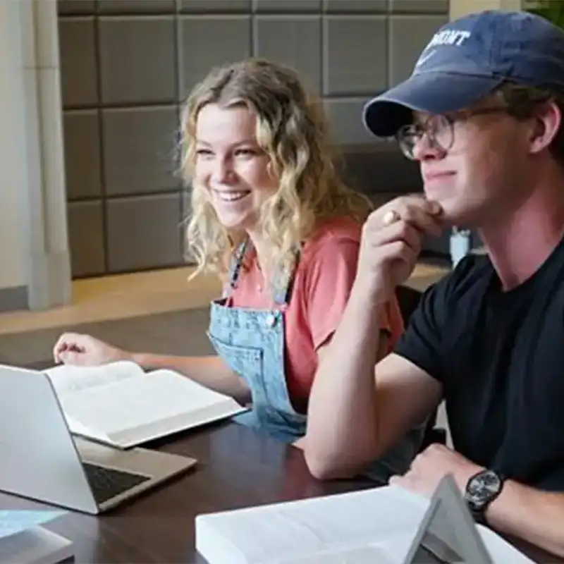 An indoor photo of a male and female seated at table with a laptop and notebook in front of them as they smile looking across the table.