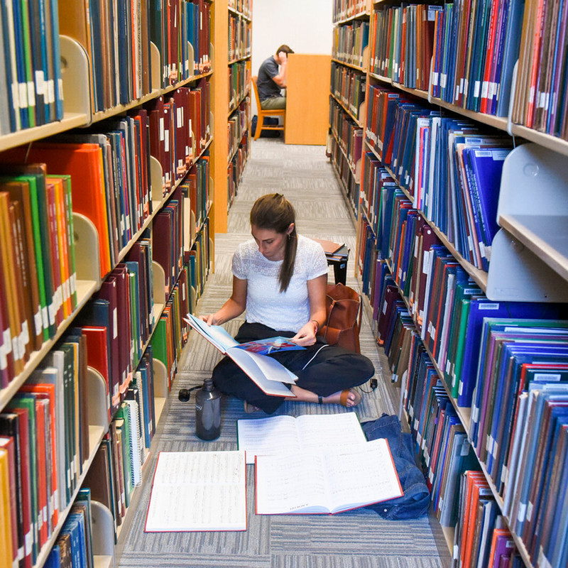 Student sitting on the floor between the bookshelves studying surrounded by books