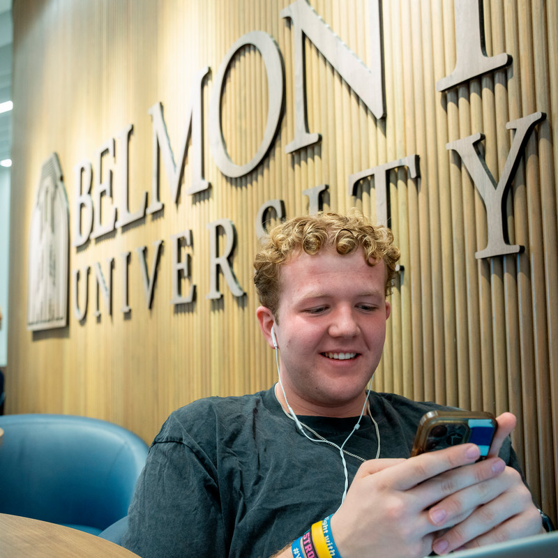 Male student sitting in front of a Belmont University sign while smiling at his phone and wearing earphones.