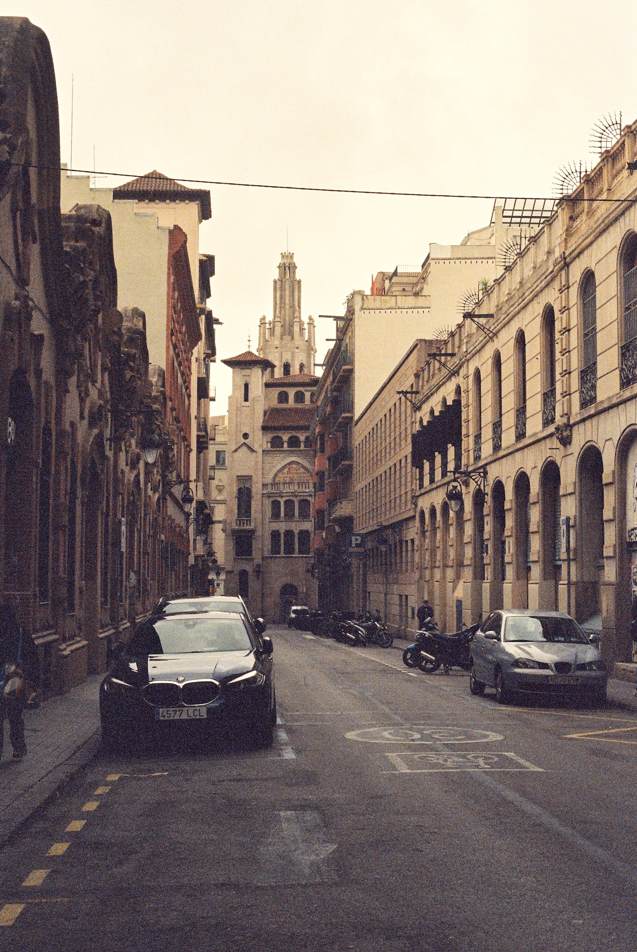 Narrow historic street in Seville, Spain, with a Gothic church tower visible at the end of the perspective. Traditional Spanish architecture lines both sides of the quiet street where cars and motorcycles are parked. The muted, vintage-toned photograph captures the charm of Seville's old town under an overcast sky.